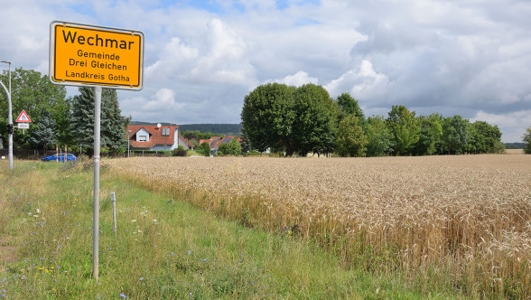 Man sieht über ein Getreidefeld auf den Ortsrand vom Bachort Wechmar. In der Bildmitte ist das gelbe Ortseingansgsschild. Dahinter sieht man Wohnhäuser, der Himmel ist wolkenlos.